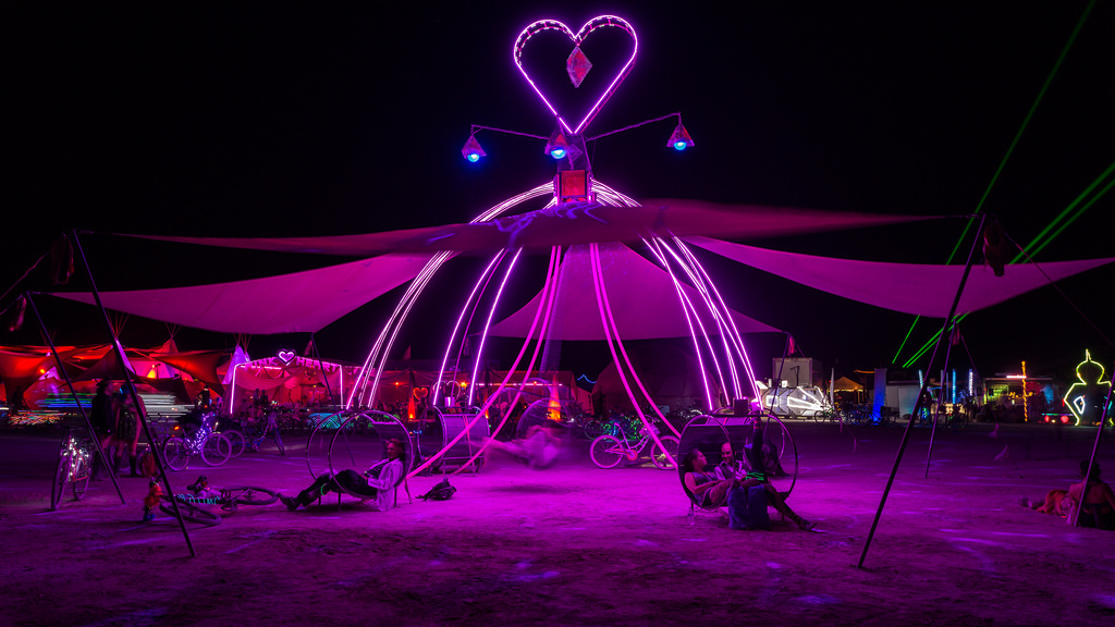 The Dream Swing at night, fully lit — arcs of pink light rising to a neon heart over swing chairs on the open playa
