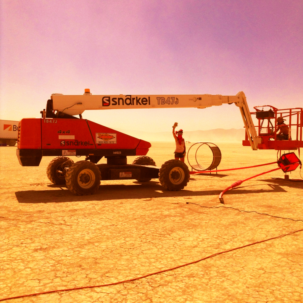A 4x4 boom lift on the open playa beside a swing chair frame, dust on the horizon