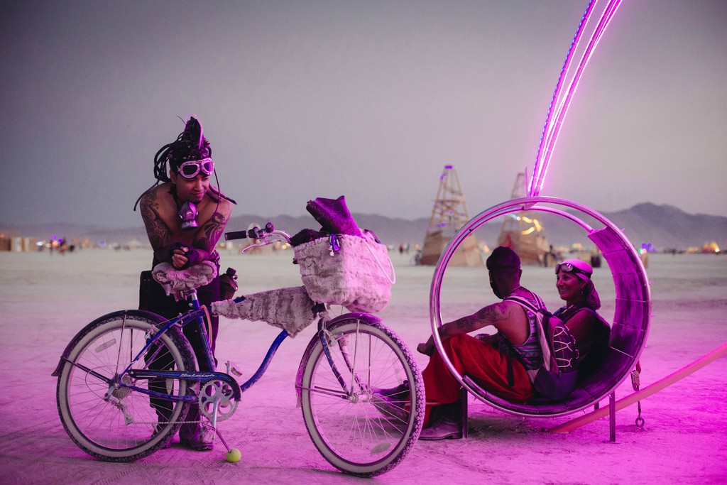 Dusk on the playa: a friend with a bike chats with two riders seated in a Dream Swing hoop chair