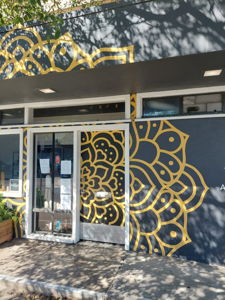 The mural seen close and square to the building: a single gold mandala continuing across the overhang fascia, down past the soffit and transom, onto the glass of the entrance door, and out across the main wall