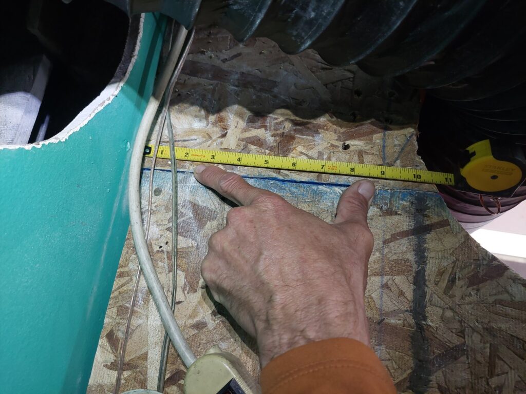 A hand holding a tape measure into an opened floor cavity, showing the depth available between the joists