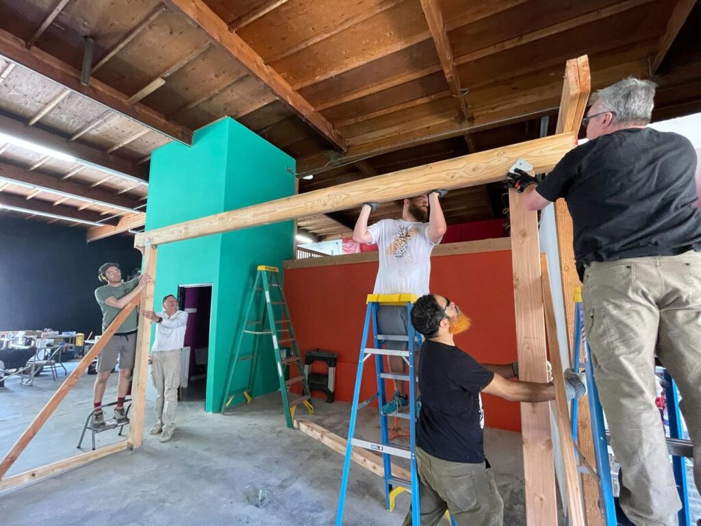 Three volunteers working together on the first framing at CoLab, one on a stepladder holding a long beam overhead against the open roof structure