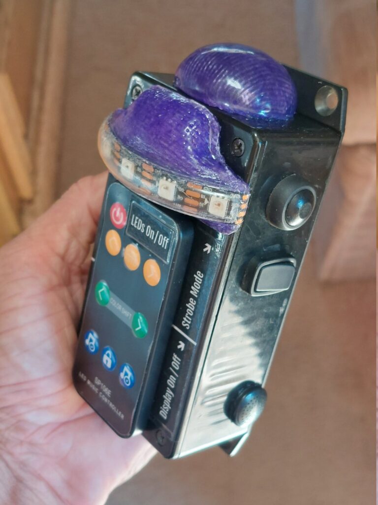 The finished controller held in one hand: an engraved face reading Display On/Off and Strobe Mode, purple potted indicator domes, and an inset LED music controller pad