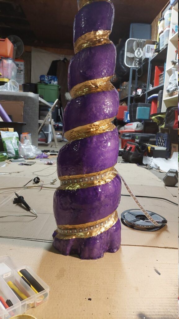 The horn standing upright on a workbench, fiberglassed and painted purple with a gold spiral, an addressable LED strip being laid into the spiral, the reel of strip alongside