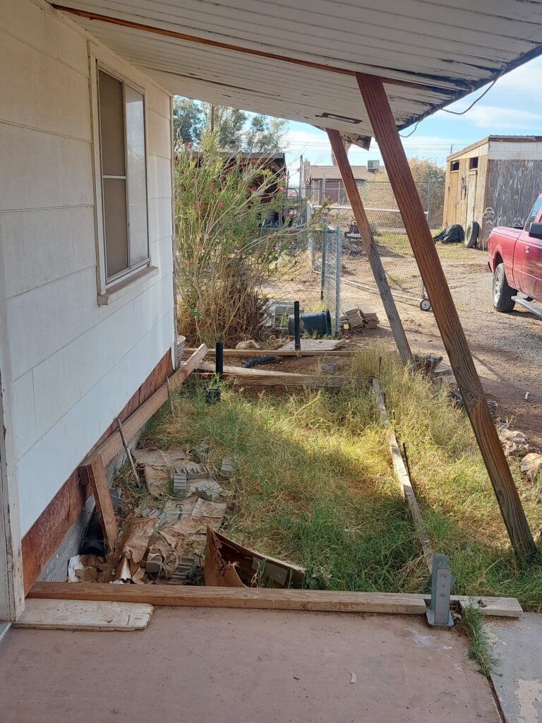 The unfinished third of a porch: grass grown up through abandoned formwork, rotted boards lying on the ground, a loose column base bracket, and the roof carried on a diagonal timber prop