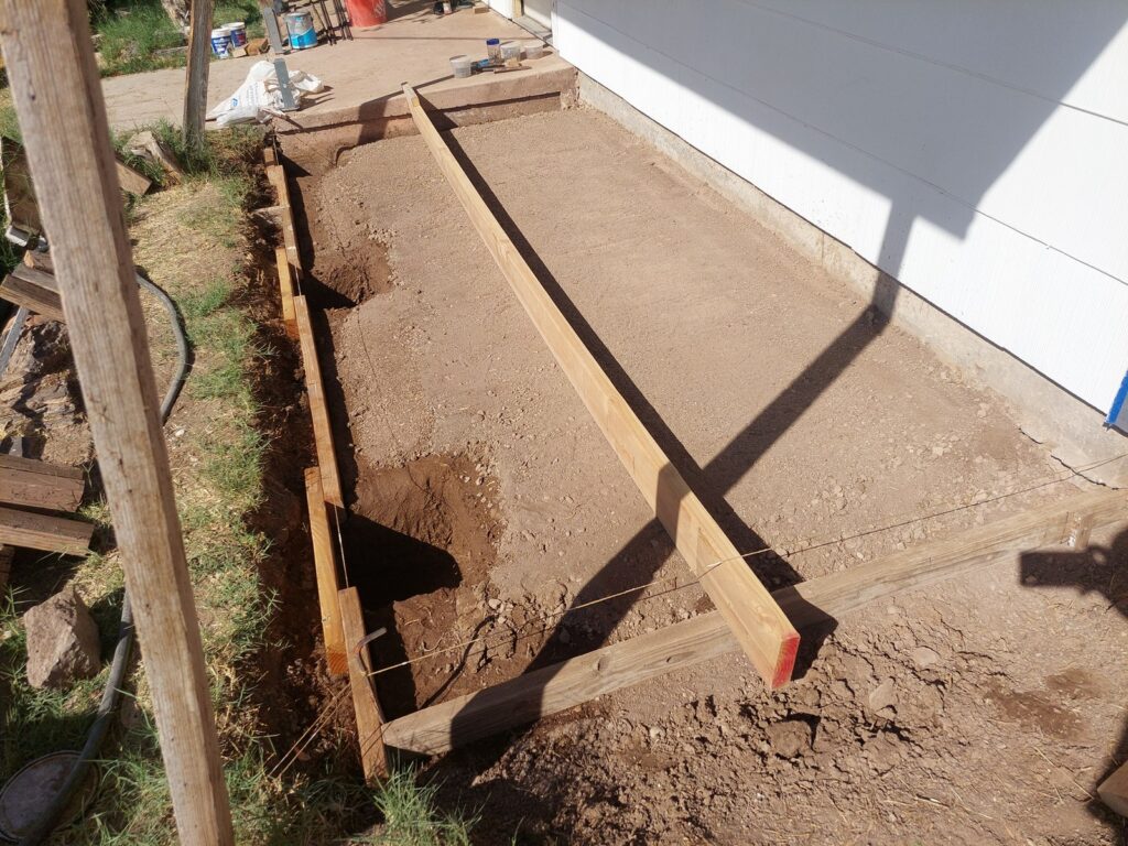 The excavated porch footprint with timber forms set along its length and a footing hole dug at one end, the house wall and existing slab alongside