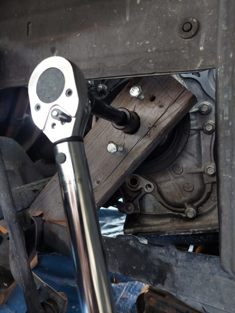 A shop-made fixture at the front of the engine: a block of wood bolted through the harmonic balancer with two bolts to stop the crankshaft turning, with a long breaker bar on the center bolt