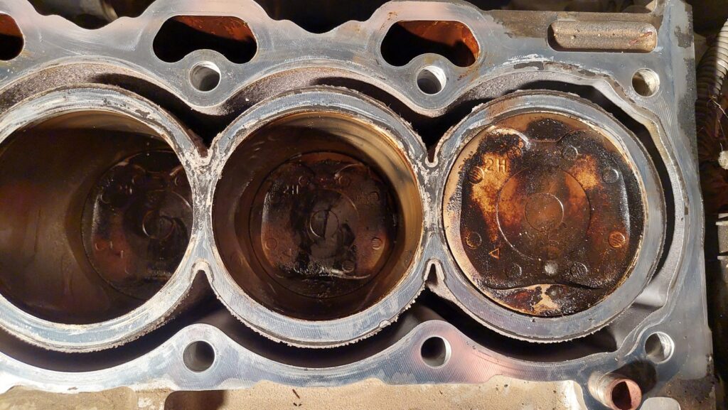 The engine block with the cylinder head removed, showing four cylinder bores. Three are dark with carbon; the second from the left is visibly clean and pale where coolant has washed the piston crown.