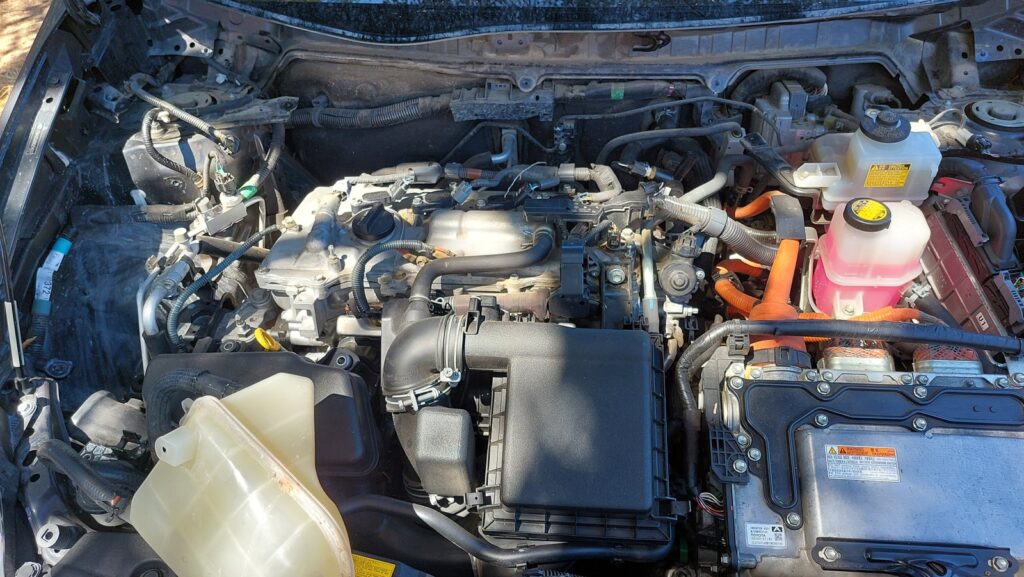 The engine bay of a 2011 Prius before any disassembly, intake and cam cover in place, high-voltage cabling visible alongside