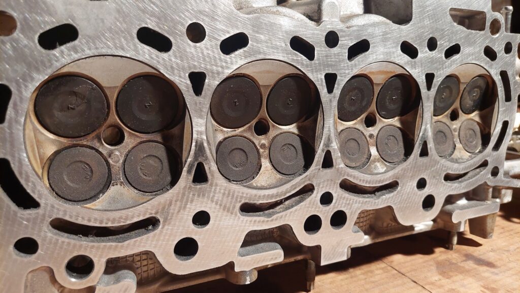 The underside of the cylinder head after cleaning, showing four combustion chambers with their valves seated and the machined sealing face around them