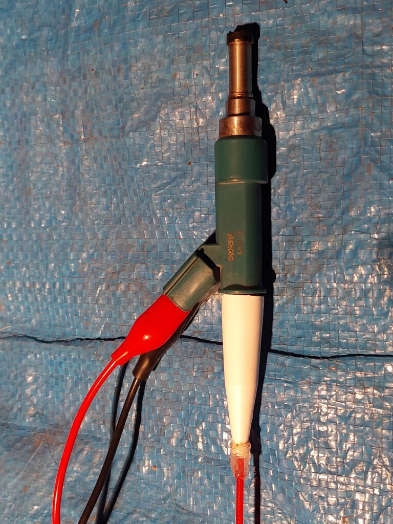 A fuel injector held in a shop-made cleaning rig: a plastic cone siliconed onto the spray tube of a cleaner can, feeding the injector, with red and black test leads clipped to its terminals to pulse it