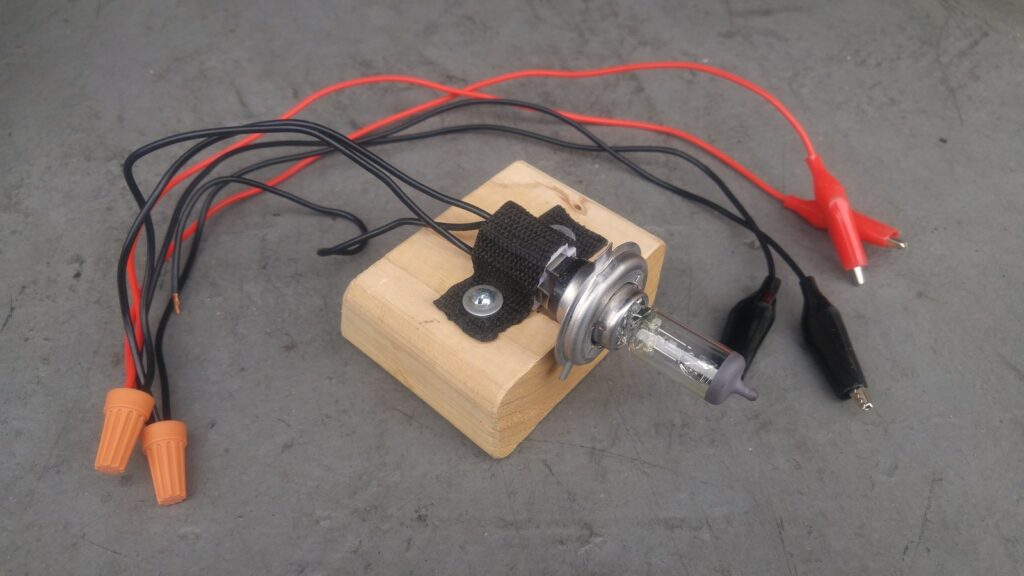 A shop-made battery load tester: a halogen headlight bulb mounted in a block of wood, wired to red and black alligator clips through wire nuts