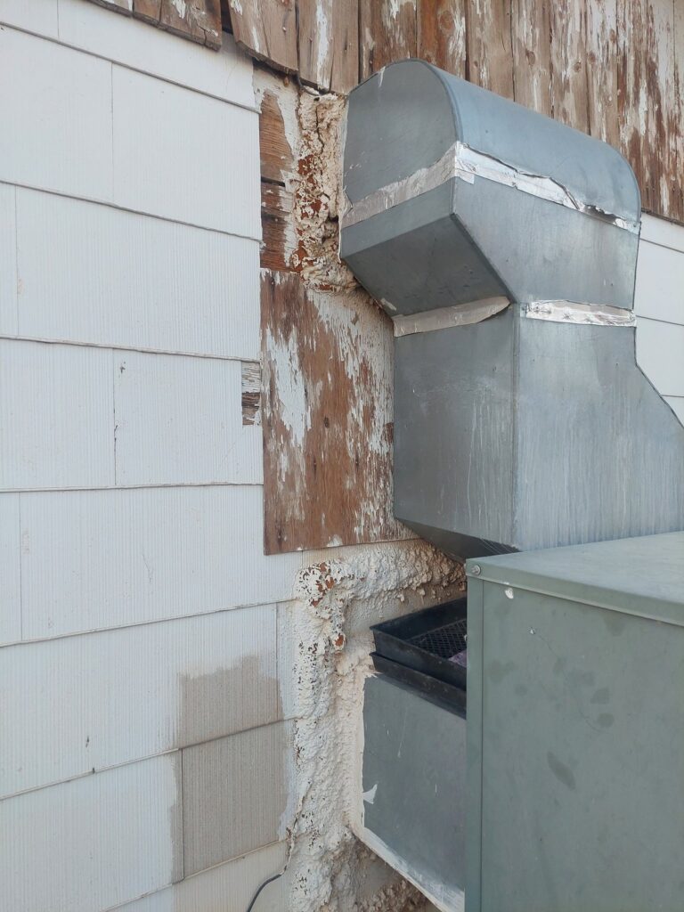An air-conditioning duct passing through the wall, the siding around it stripped and the sheathing weathered and rotted