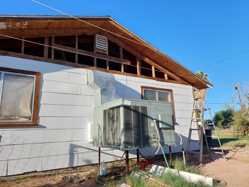 The house mid-job: the gable end stripped back to bare rafters, siding part replaced, a ladder against the wall and the air-conditioning unit in the foreground