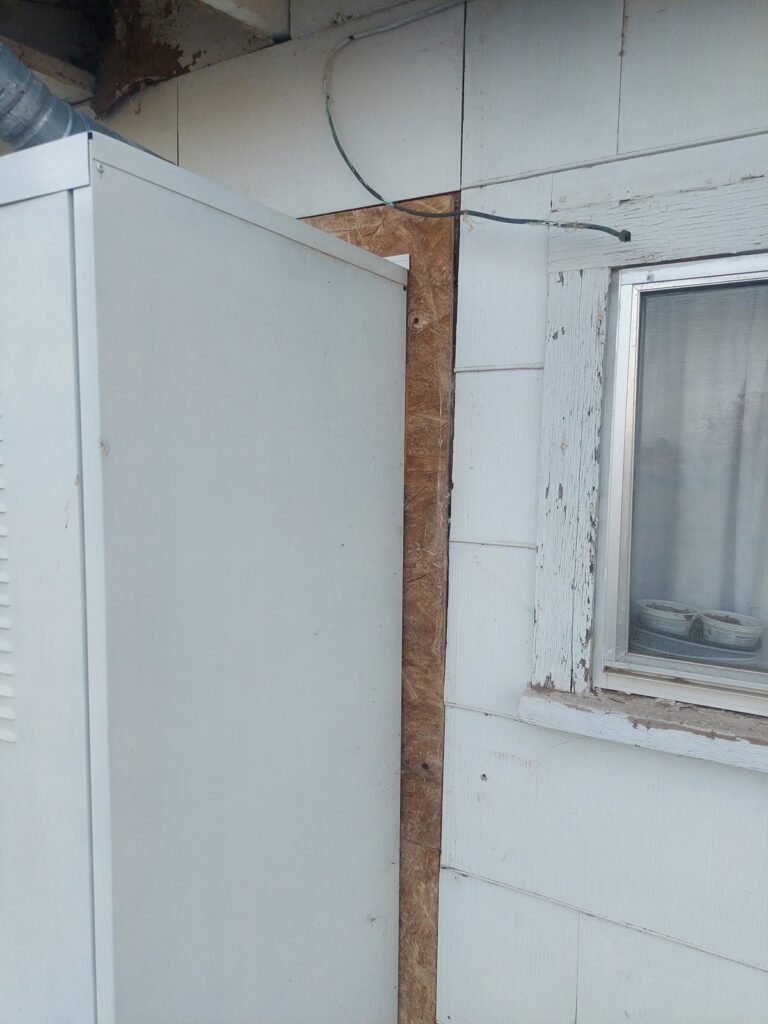 The wall beside the water-heater enclosure with siding removed, bare sheathing and an oriented-strand-board patch visible next to the window