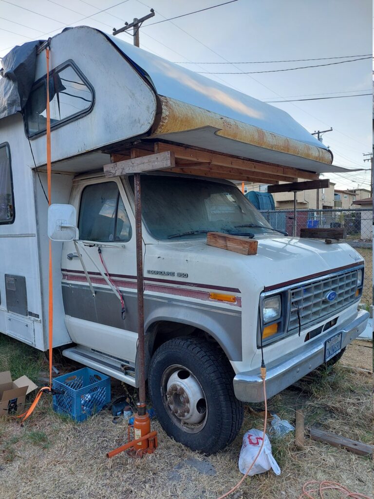 A Class C motorhome on jack stands, its cabover skin lifted and propped open on posts