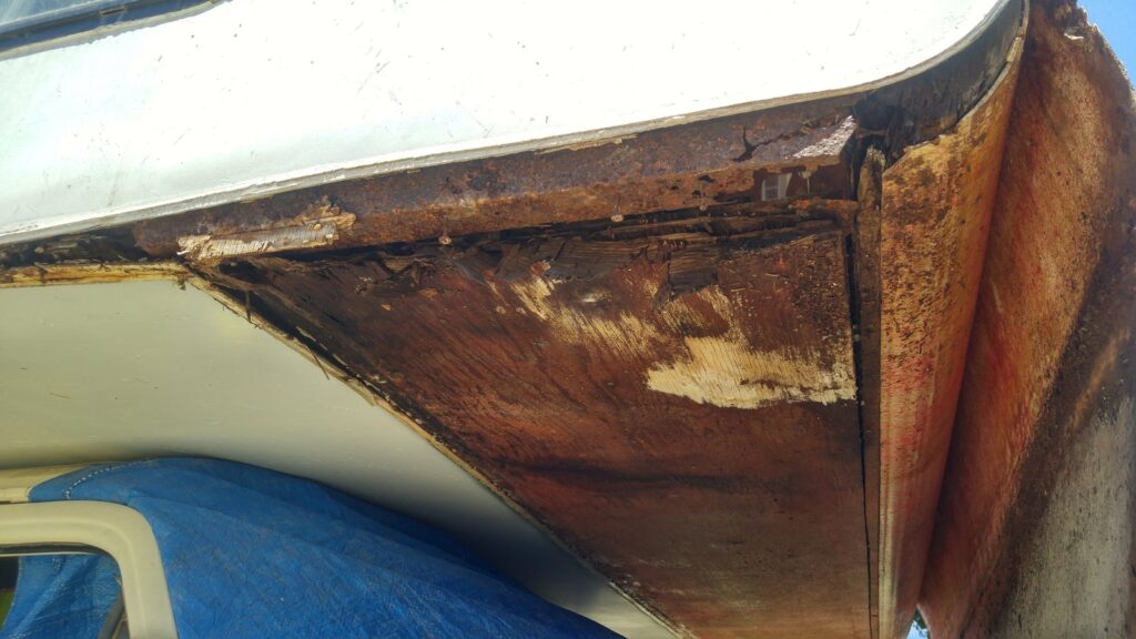 Steel tubing above the RV front window, rusted through, with rotted wood behind the peeled-back panel