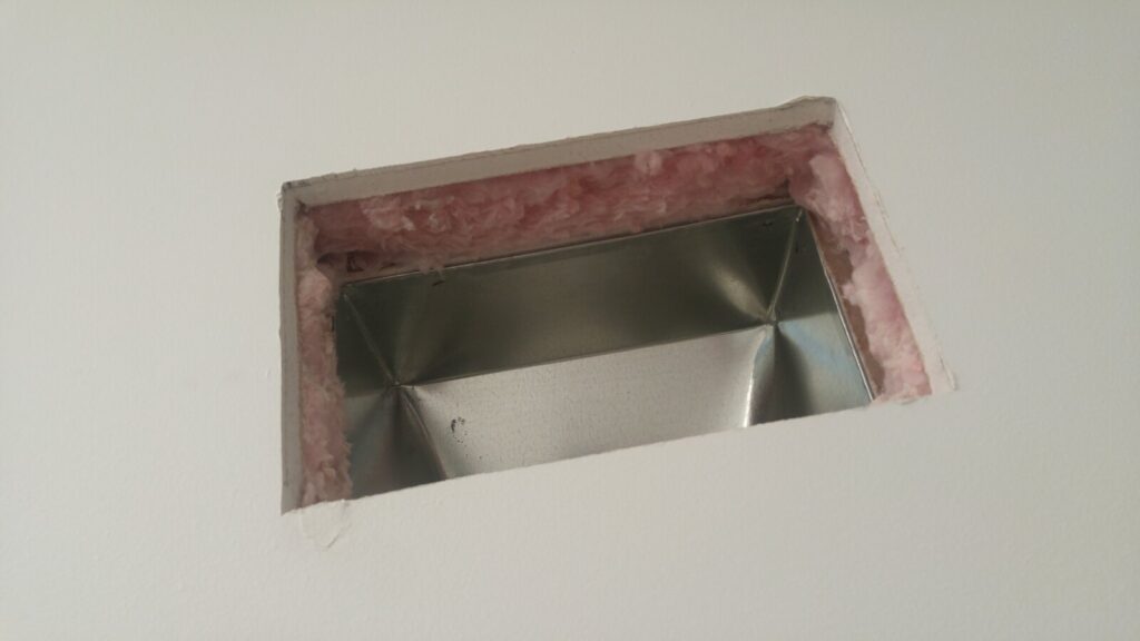 A rectangular opening cut in a wall, pink fibreglass insulation visible around its edge, with a hand-formed sheet-metal boot fitted inside it