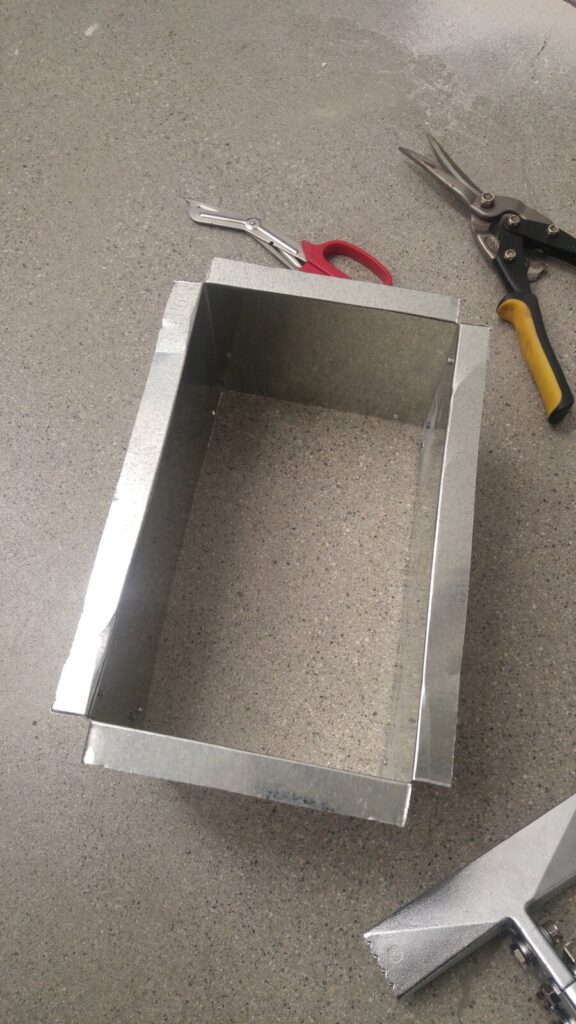 A rectangular sheet-metal duct box formed by hand, resting on a concrete floor beside aviation snips and a hand seamer