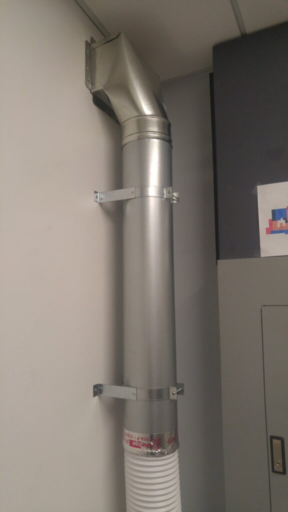 A rigid round metal duct running up a wall to an elbow near the ceiling, held by two wall straps