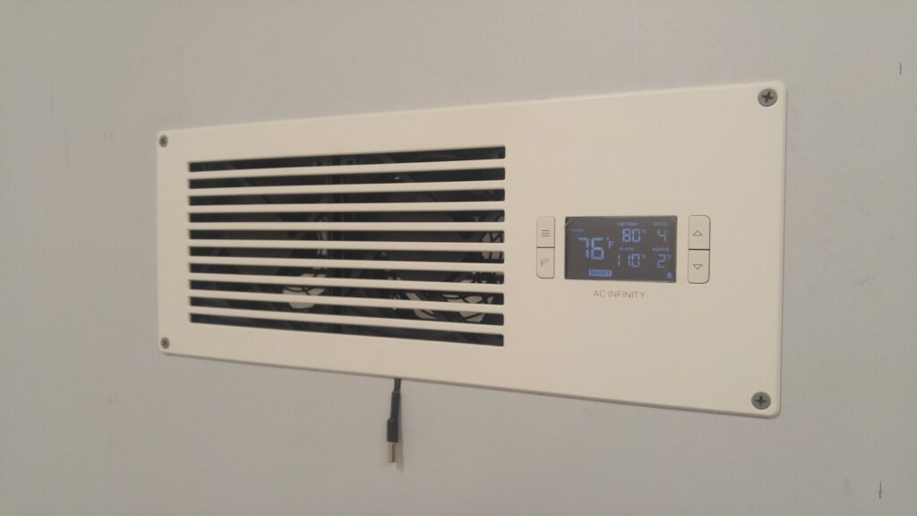 A white louvered fan unit set into a wall, with a small digital thermostat display on its face showing a temperature setting