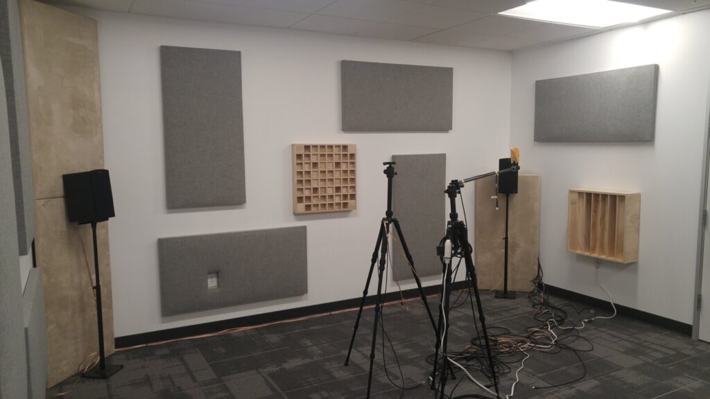 A small room with grey absorber panels and slotted wood diffusers on white walls, a bass trap in the corner, monitor speakers on stands and two measurement tripods on the floor